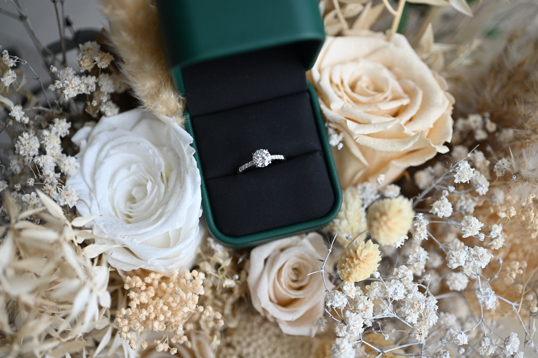 Elegant inel de logodnă din platina cu diamant 1.01ct și diamante lab grown, prezentat într-o cutie verde, înconjurat de flori albe.